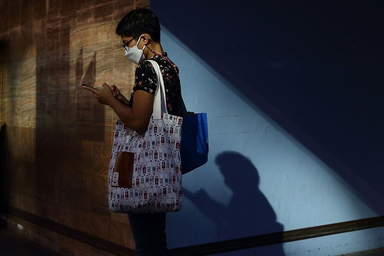 シンガポールでバスを待つ間、携帯端末を使用するマスク姿の女性/Suhaimi Abdullah/NurPhoto/Shutterstock