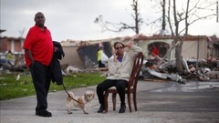 破壊された自宅の横の通りで椅子に座り電話をかける女性