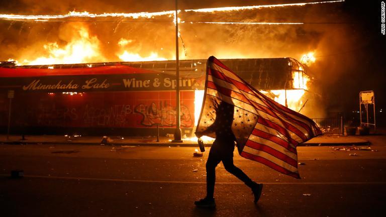 星条旗を逆さまに構え、燃え上がる建物の前を進むデモ参加者/Julio Cortez/AP