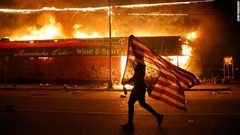 星条旗を逆さまに構え、燃え上がる建物の前を進むデモ参加者