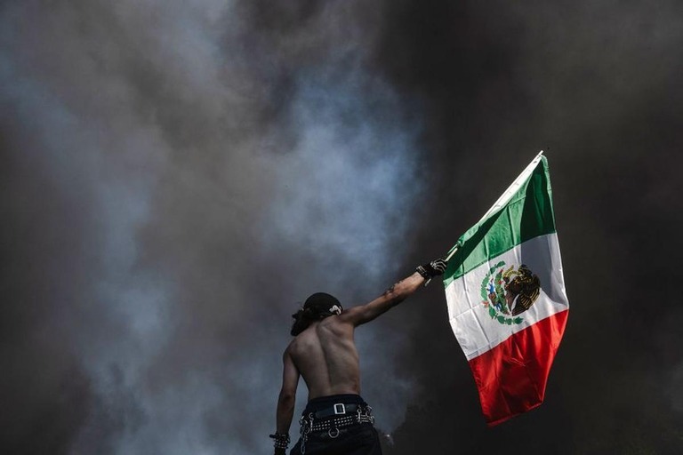 ８日、燃える車両から黒煙が立ち上る中でメキシコ国旗を掲げる抗議デモ参加者/Jim Vondruska/Getty Images