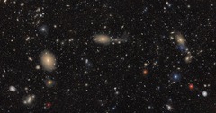 地球から約５５００万光年離れたおとめ座銀河団。赤や青に輝く星のほか、近くの青い渦巻き銀河や遠方の赤い銀河群も見える