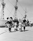 ディズニーランドの開園当日、ミッキーマウスとミニーマウスが海賊船のそばで子どもたちと一緒に歩く様子